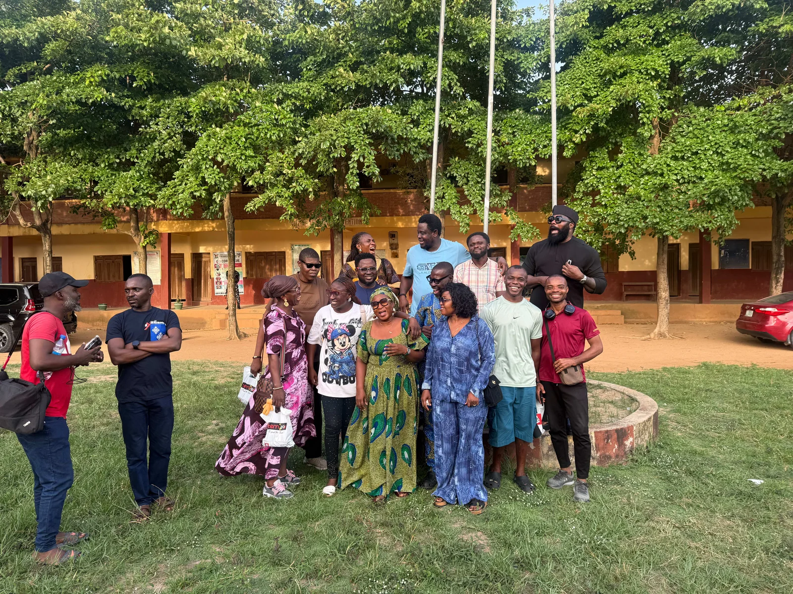 Wide view of FGSCAA members laughing together on the lawn of Festac Grammar School, with tall trees, flagpoles and the classroom block behind them.