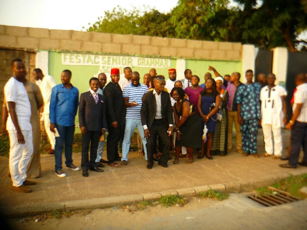 A large group of members standing on the pavement in front of the school wall painted “Festac Senior Grammar School”, one pointing towards the school.