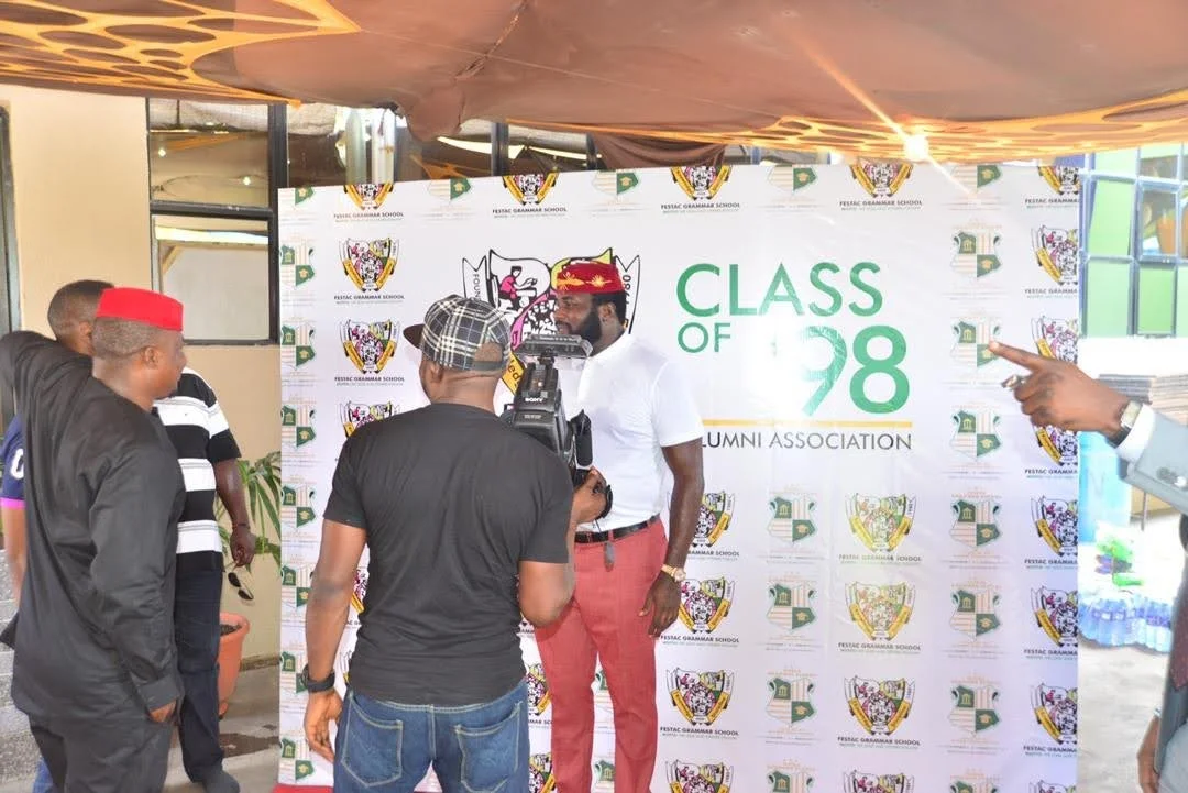 A camera operator filming a member standing before the Class of 98 Alumni Association backdrop while other members look on.