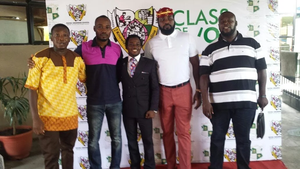 Five members standing side by side in front of the Festac Grammar School crest and Class of 1998 backdrop.