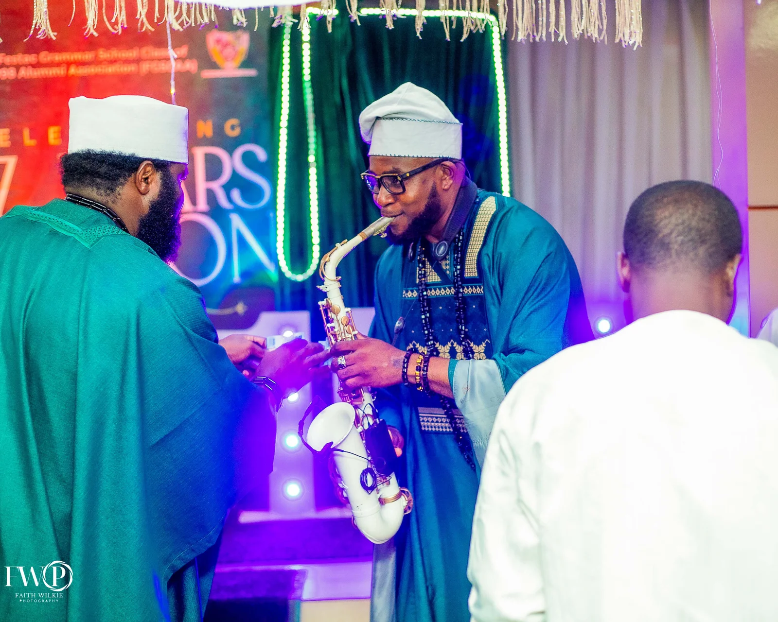 A saxophonist performing live in front of the reunion backdrop while a member in green attire appreciates the performance.