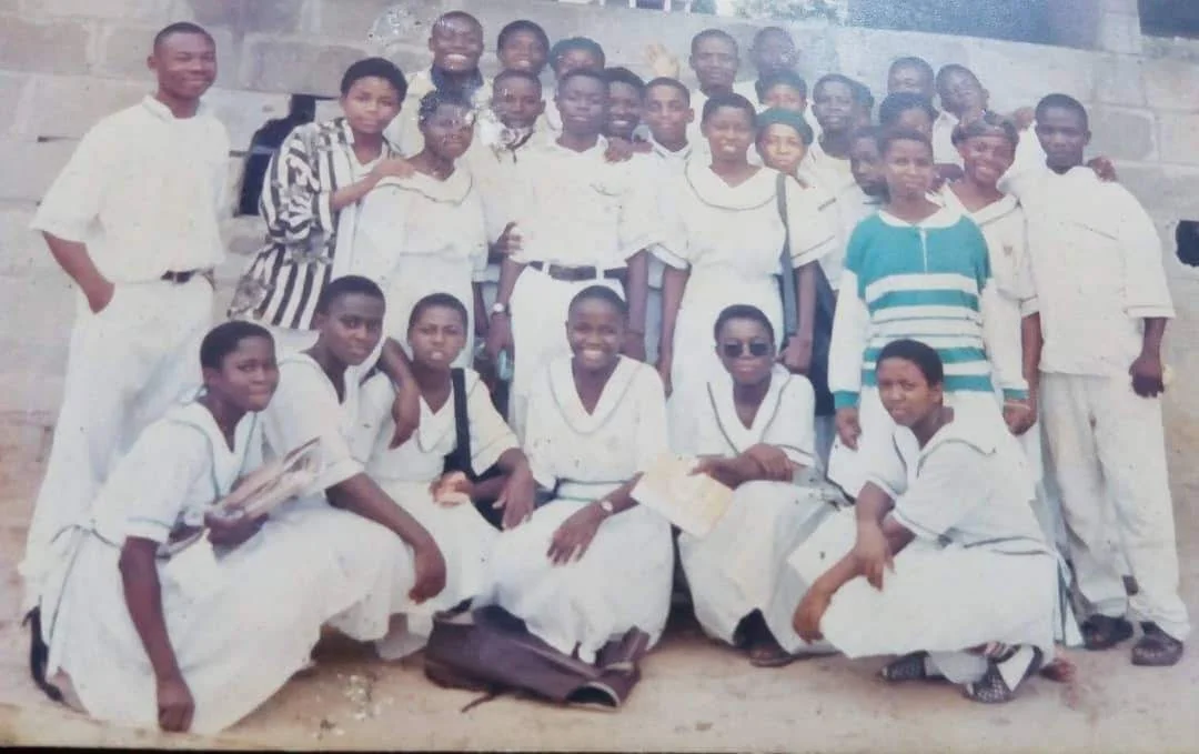 A large group of Festac Grammar School students in white school uniform, some seated in front and others standing behind, posed against a block wall.