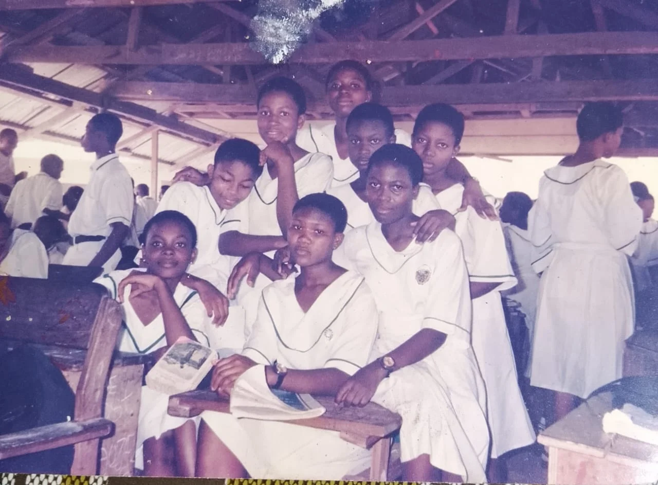 Eight female students in white uniforms gathered around wooden desks beneath an open timber roof, with other students in the background.