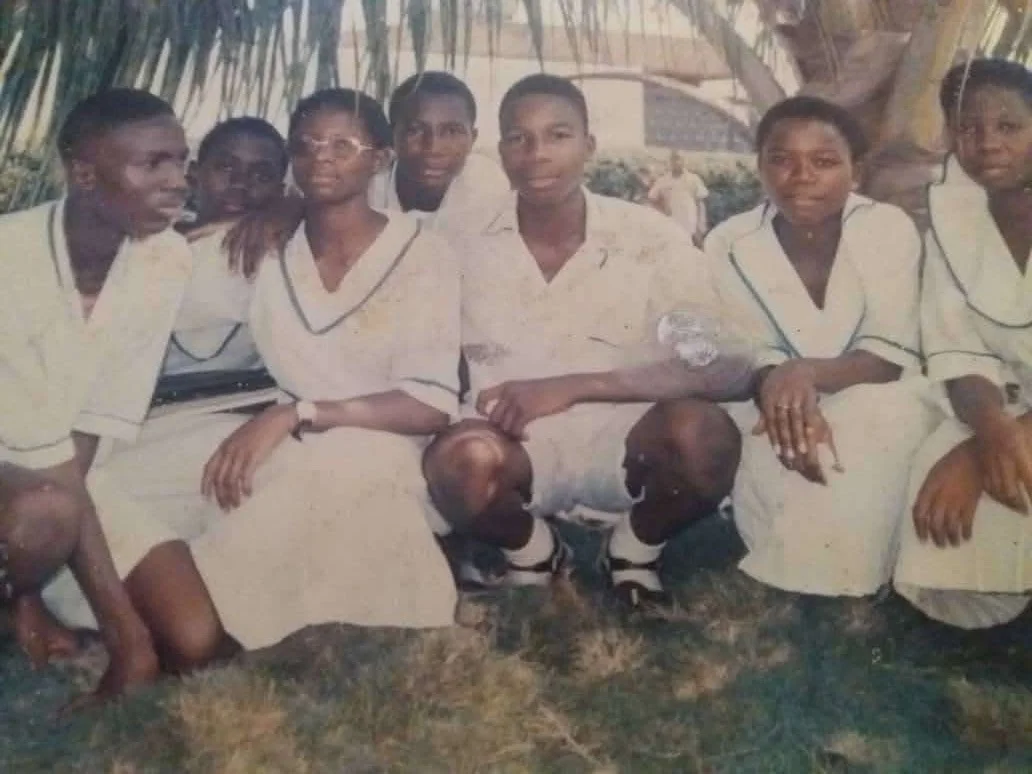 Seven students in white uniforms crouching and kneeling together on grass beneath a palm tree.