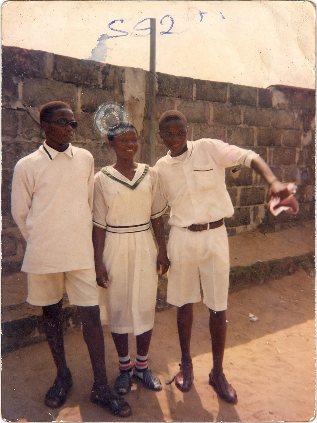 Two male students in white shirts and shorts standing either side of a female student in a white uniform with green trim, in front of a block wall.