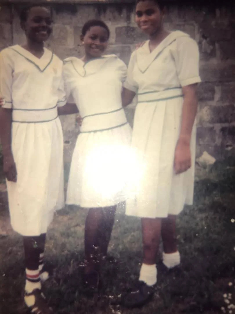 Three female students in white uniforms with green trim standing arm in arm on grass in front of a block wall.