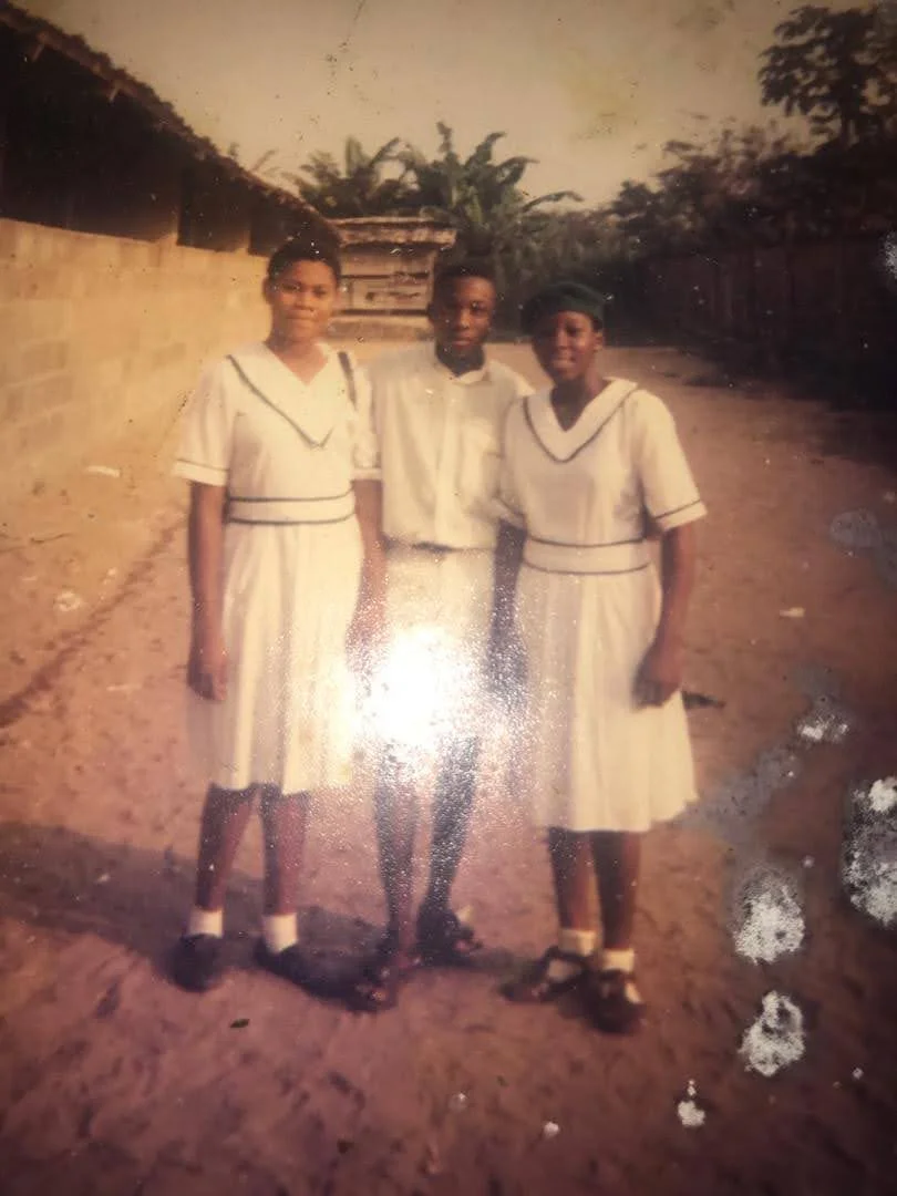 A male student between two female students in white uniforms, standing on a sandy path beside a long block building.