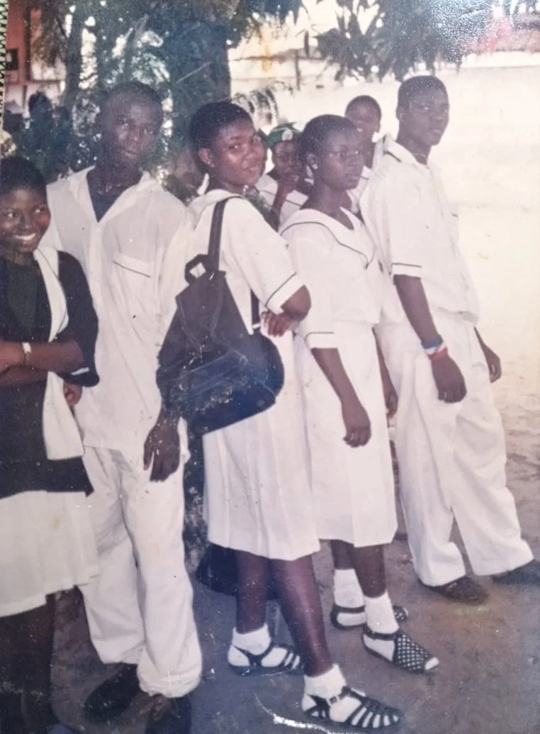 A line of students in white uniforms standing together outdoors, one carrying a dark backpack.