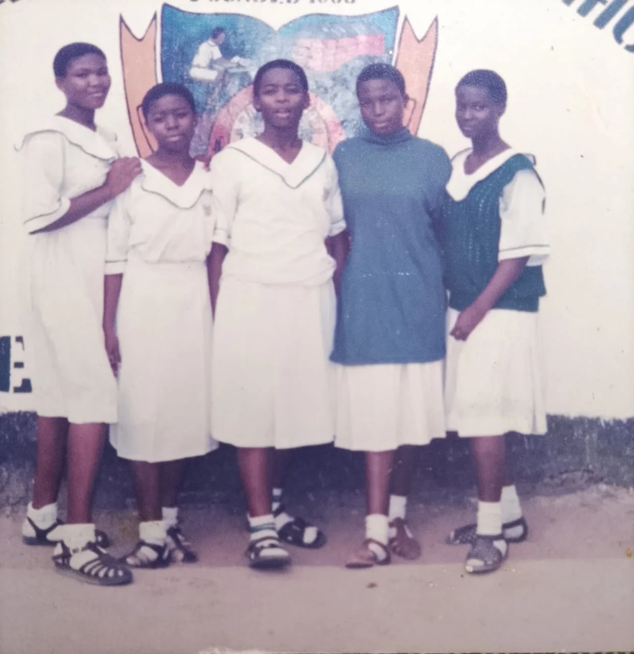 Five female students, two wearing green sweaters over their white uniforms, standing in front of a painted Festac Grammar School crest on a wall.