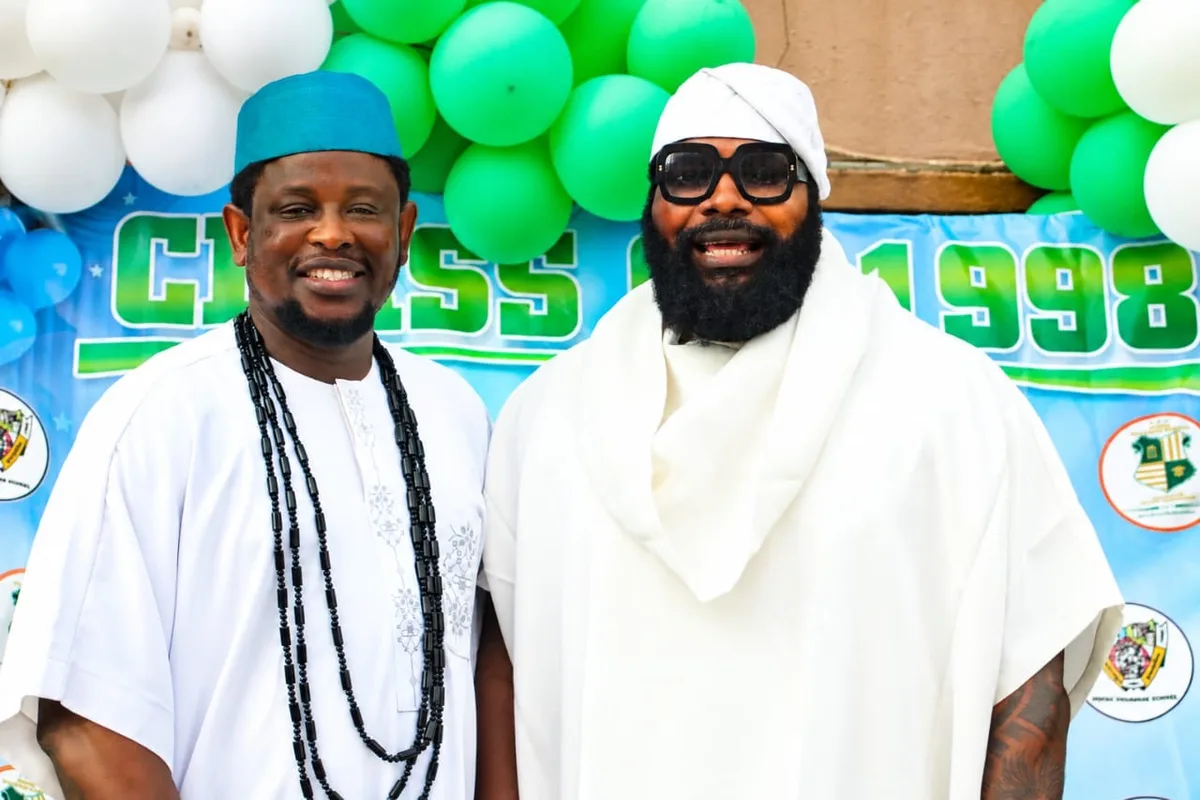 Two members in white attire smiling in front of a Class of 1998 banner and balloons