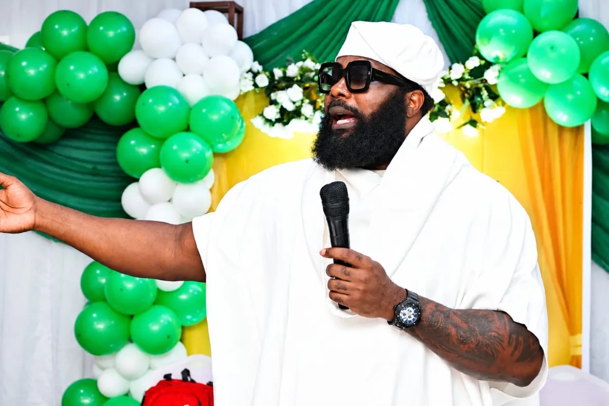 A member in white attire speaking into a microphone with one arm outstretched, in front of green and white balloons