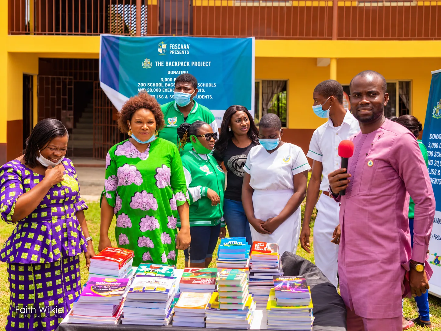 FGSCAA members and students beside a table of donated books under a Backpack Project banner, as a speaker addresses the event.