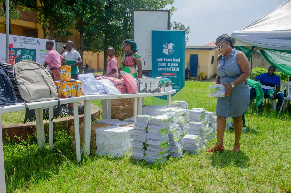 Close view of school bags, boxes of pens, uniforms and stacked notebooks on tables and the grass, with an FGSCAA member carrying a bundle of notebooks.