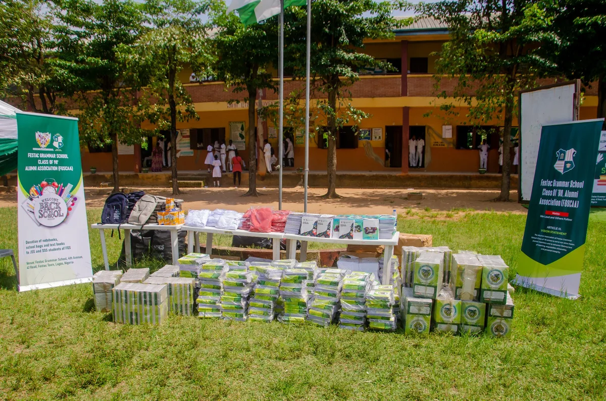 Notebooks, school bags, pens and uniforms laid out on tables and the grass in front of a Festac Grammar School building, between two FGSCAA Back-to-School banners.