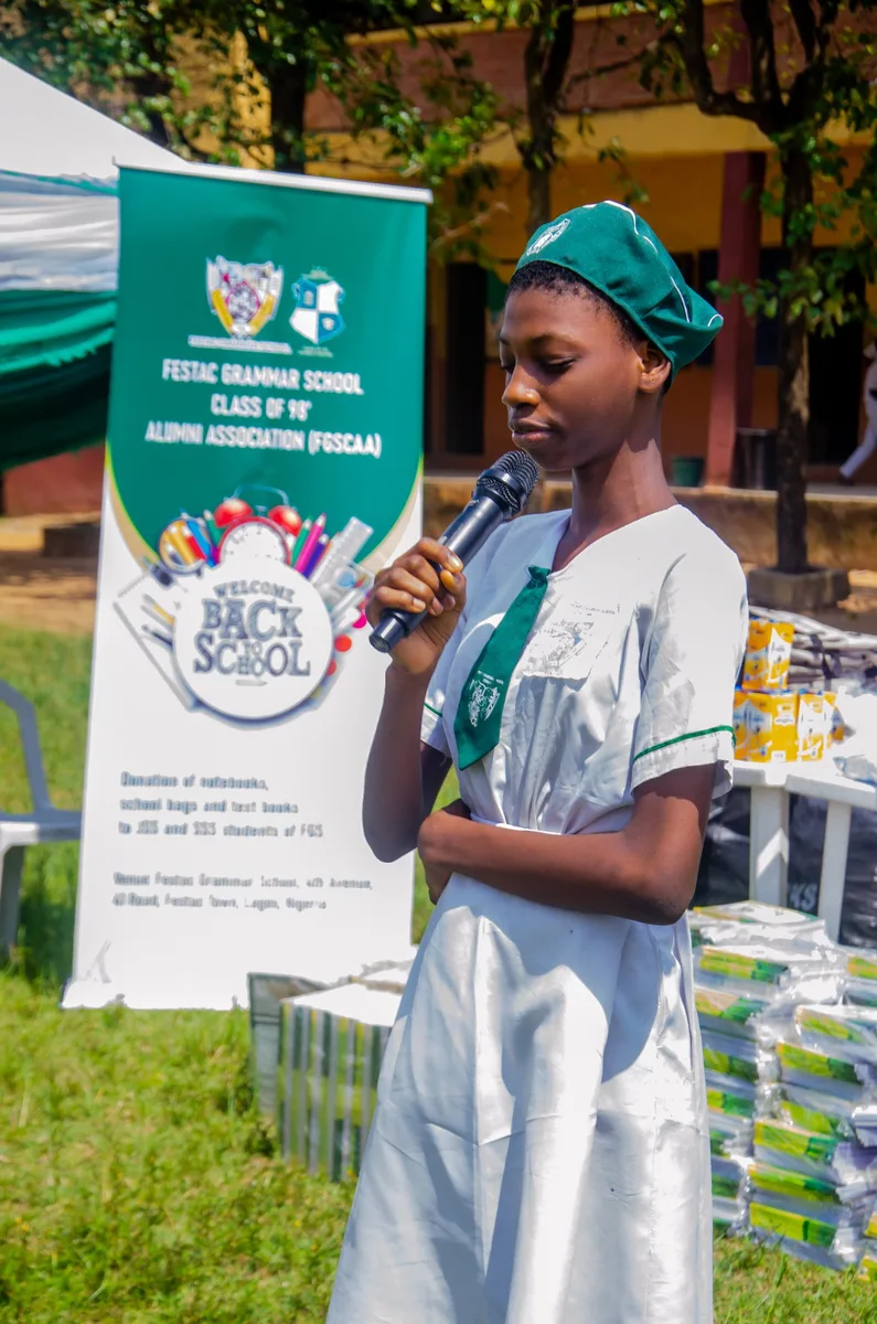A Festac Grammar School student in a green beret and white uniform speaks into a microphone, with an FGSCAA Back-to-School banner and donated notebooks behind her.