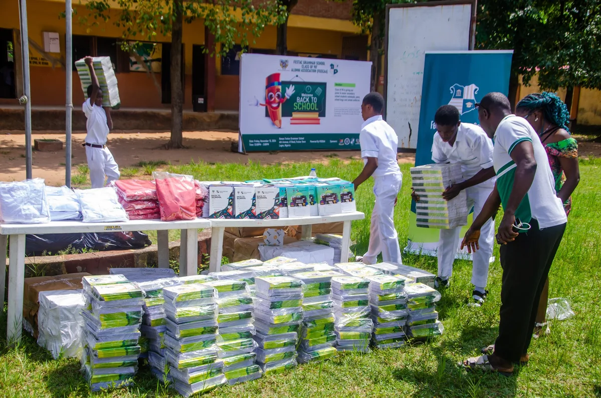 Students in white uniforms carry bundles of wrapped notebooks across the field while an FGSCAA member looks on beside stacks of donated materials.