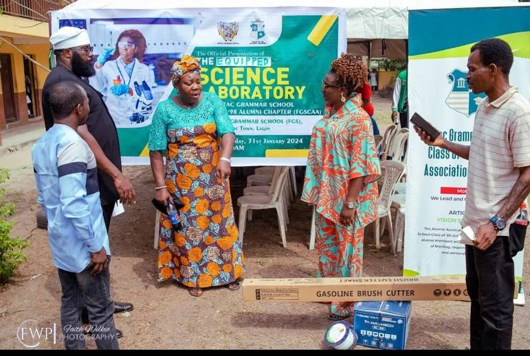 Members and school representatives stand around a boxed gasoline brush cutter and Maxmech equipment box displayed on the ground.
