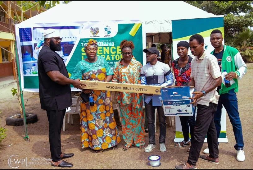 An FGSCAA representative in black hands a boxed gasoline brush cutter to a school representative in a patterned dress, as others hold a Maxmech equipment box.