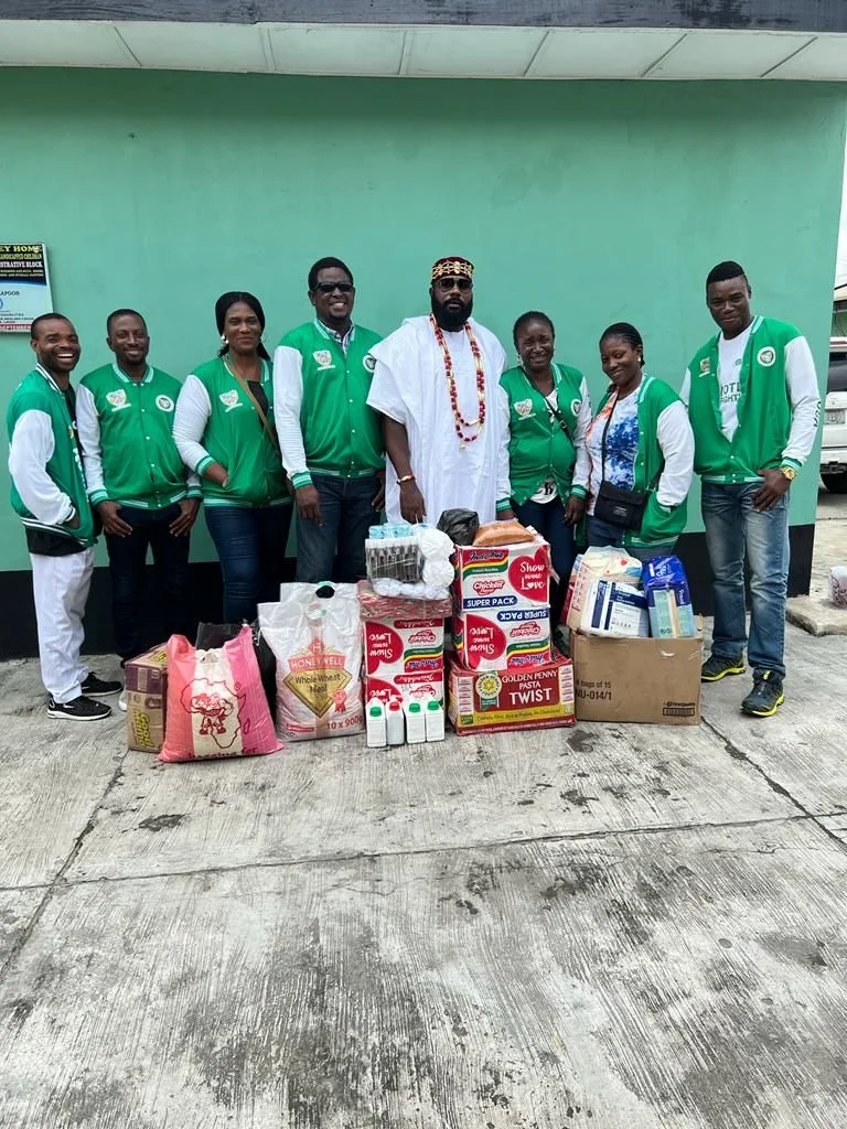 Nine FGSCAA members, most in green Association jackets, stand outside a green building behind cartons of noodles and pasta, bags of flour, liquid soap and other relief items.