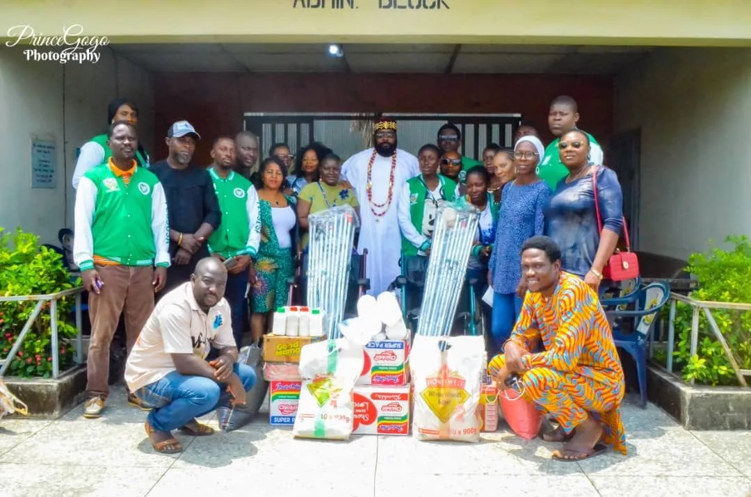 A large group of FGSCAA members and participants gathers at the entrance of an Admin Block with wrapped crutches, a wheelchair, food cartons and bags of wheat meal.