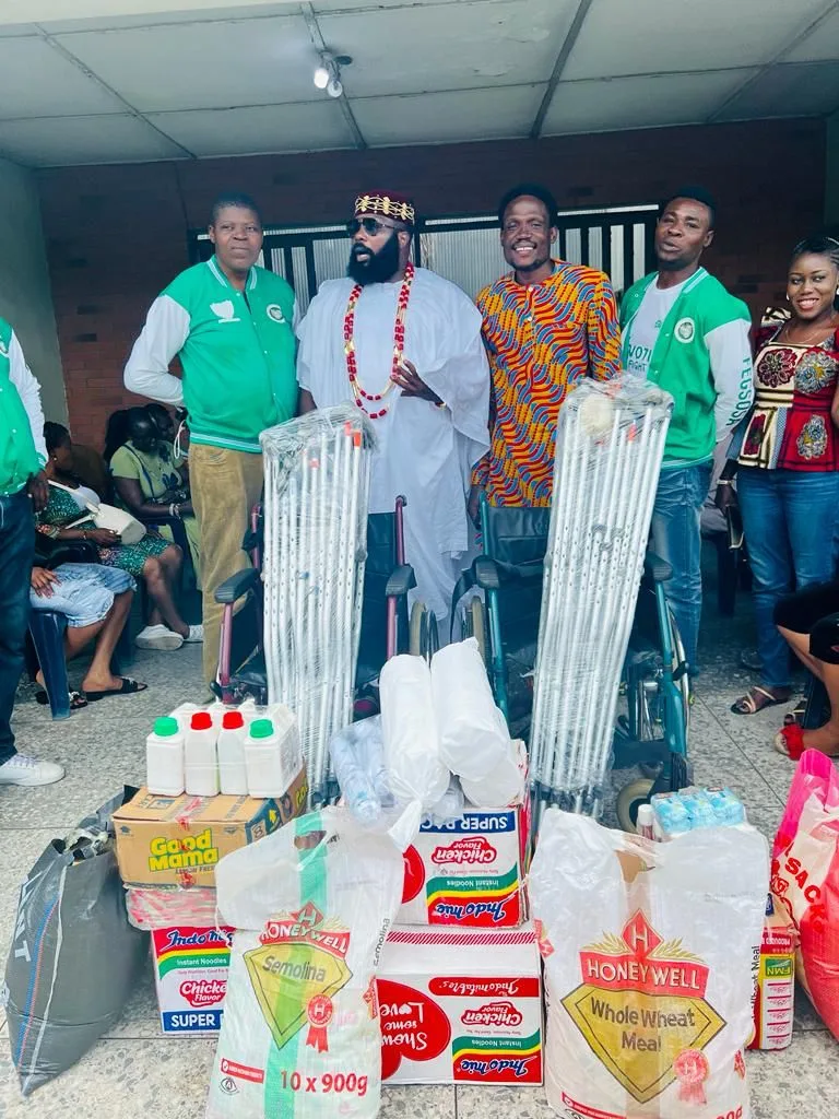 FGSCAA members and hosts stand indoors behind two wheelchairs and bundles of crutches, with cartons of noodles, bags of semolina and wheat meal and liquid soap in front.