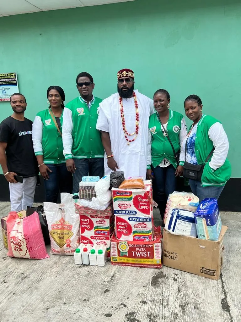 Seven members of the Association stand against a green wall behind donated cartons of noodles and pasta, bags of wheat meal, liquid soap and boxed essentials.