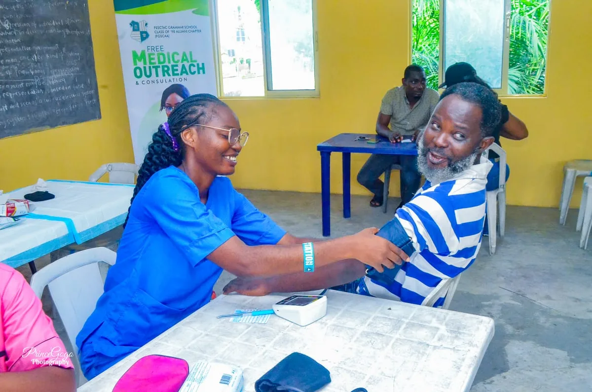 A health worker in blue scrubs fits a blood-pressure cuff on a smiling man in a striped shirt, with the FGSCAA Free Medical Outreach banner behind.