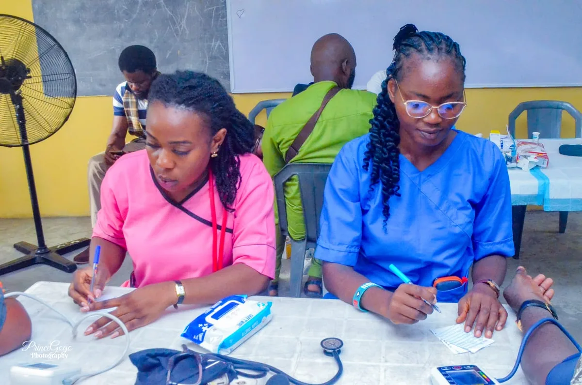 Two health workers in pink and blue scrubs record readings at a table with blood-pressure monitors and a stethoscope.