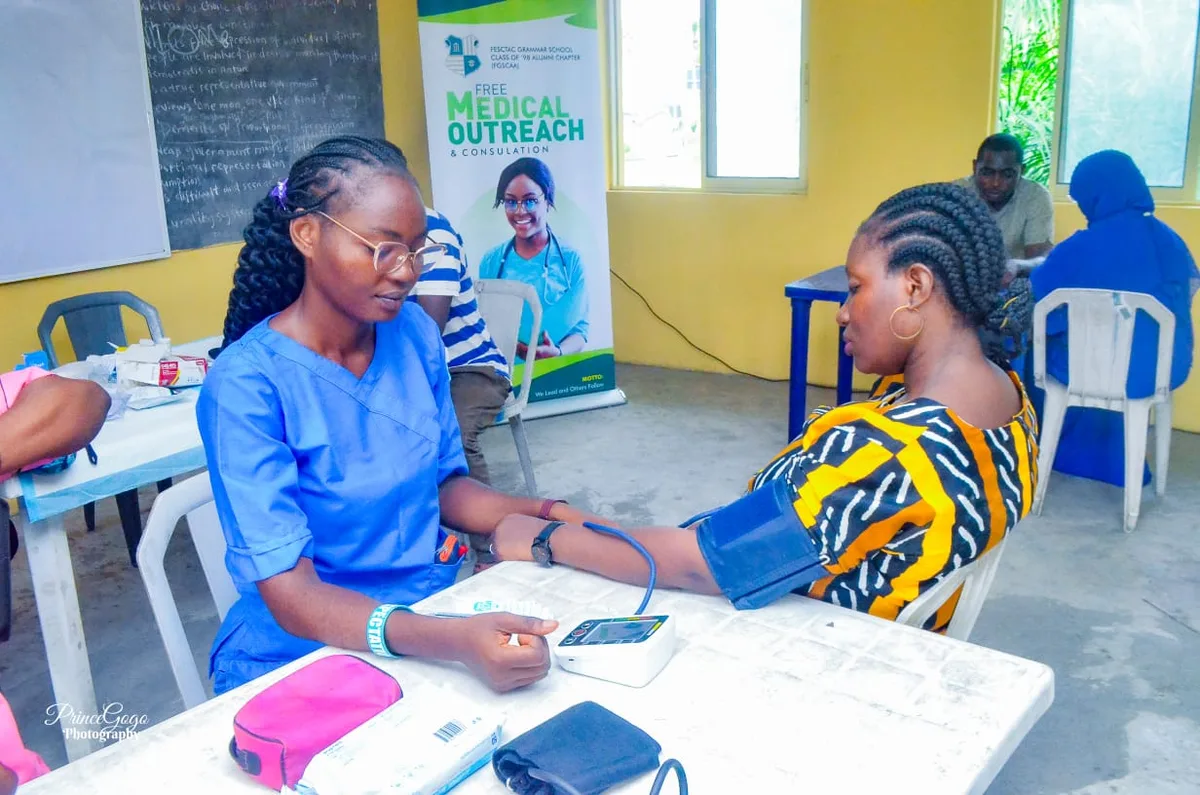 A health worker takes a blood-pressure reading beside a table of medical supplies, including a digital monitor, cuff and wipes.