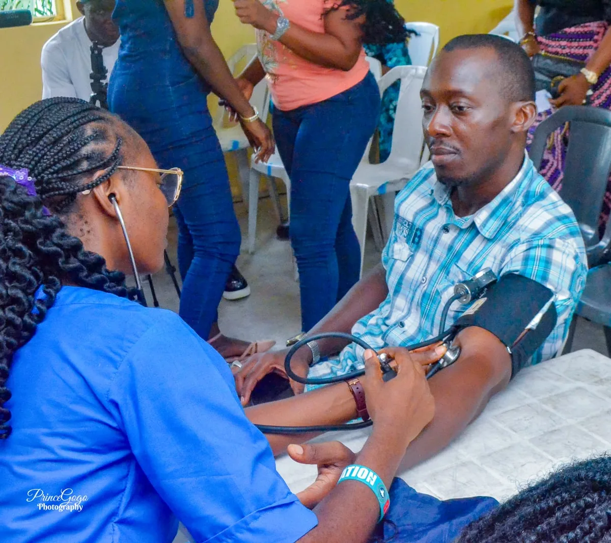A health worker uses a stethoscope and manual cuff to check the blood pressure of a seated man in a checked shirt.