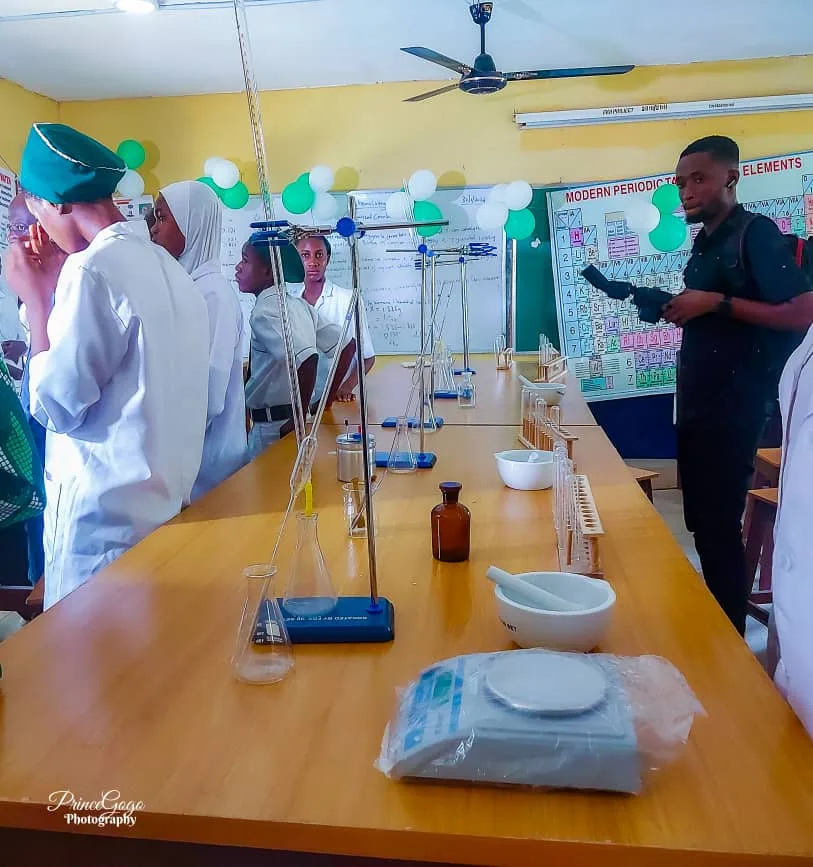 Laboratory workbenches set with retort stands, flasks, mortars and a weighing balance, with students in lab coats and a periodic table on the wall.