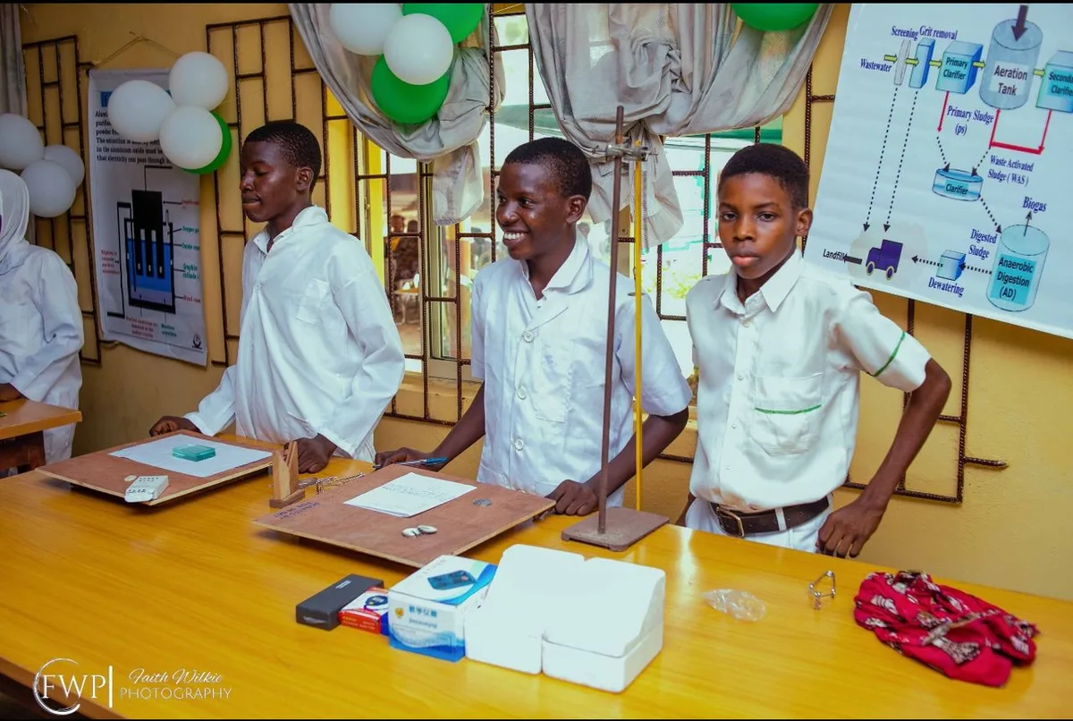 Three students in white lab coats standing at a workbench with practical apparatus and worksheets.