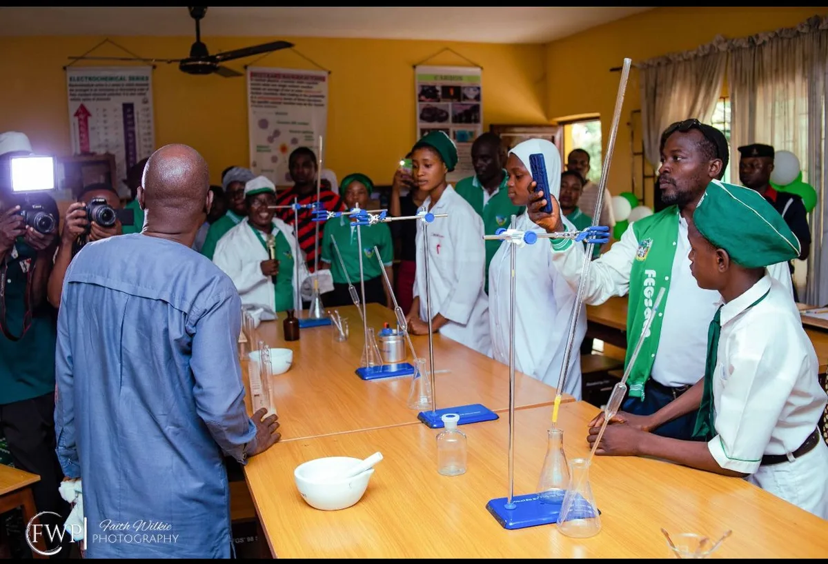 Students in lab coats beside workbenches fitted with retort stands and burettes as guests look on.