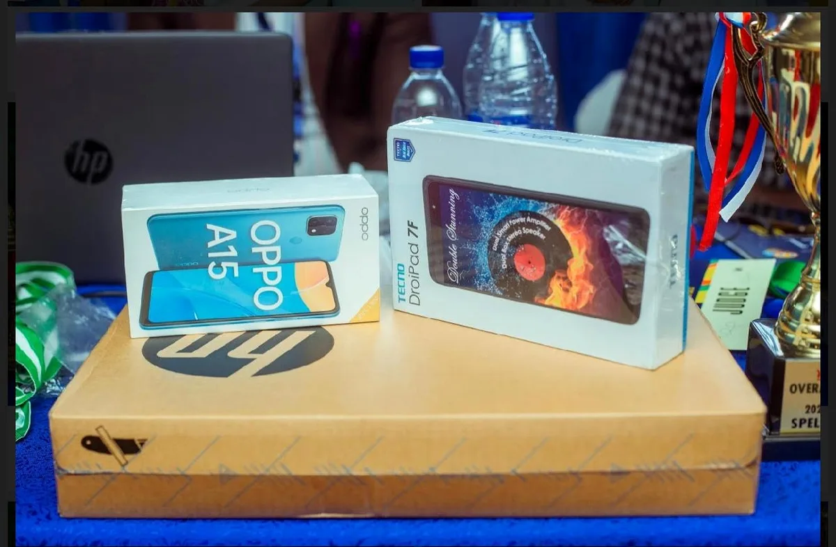 Boxed laptop, mobile phone and tablet prizes displayed beside medals and a trophy for the 2021 Spelling Bee winners.