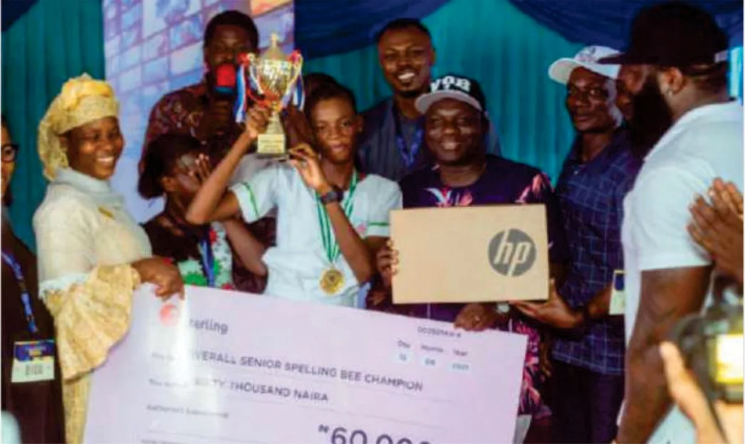 The Overall Senior Spelling Bee Champion raising a trophy, with a laptop prize and prize cheque presented.