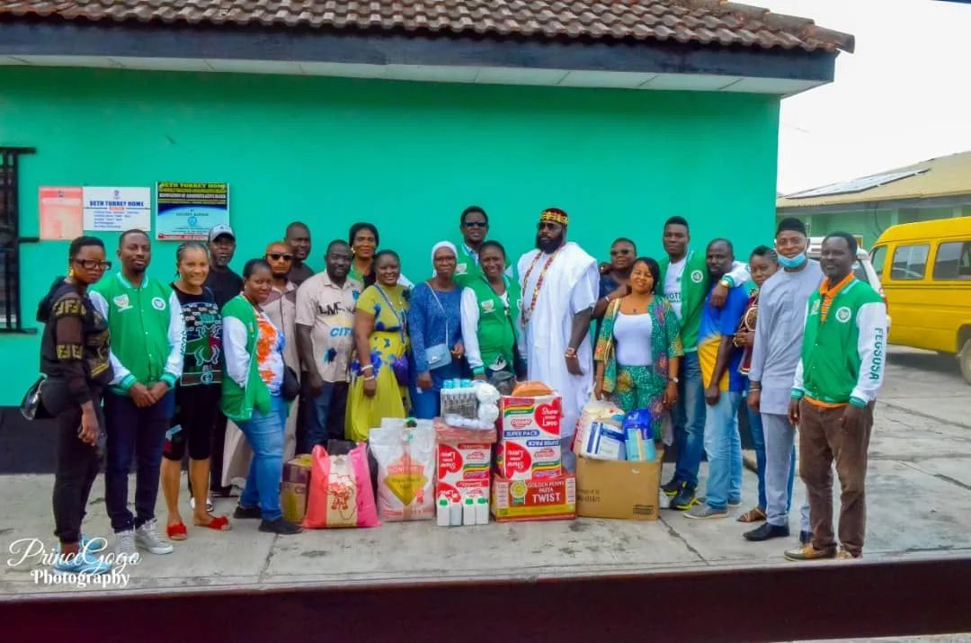 A large group of FGSCAA members and participants stands outside a green building behind donated food cartons, bags of flour and semolina, and household supplies during the 2022 humanitarian outreach.